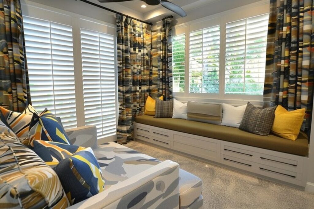 Cozy window-seat area with built-in storage drawers, white shutters, and colorful curtains; yellow, gray, and blue cushions.